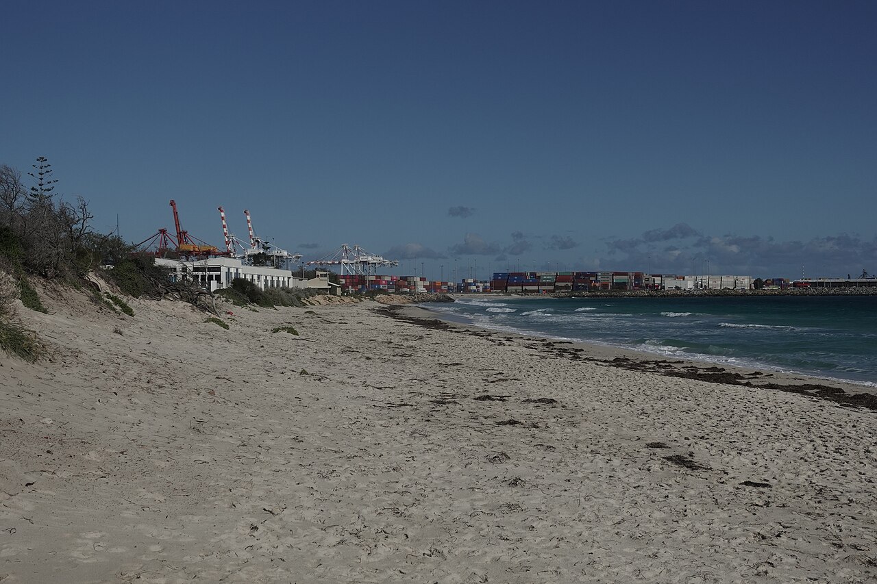 Sand and sea at Port Beach with port cranes and container stacks in the distance.