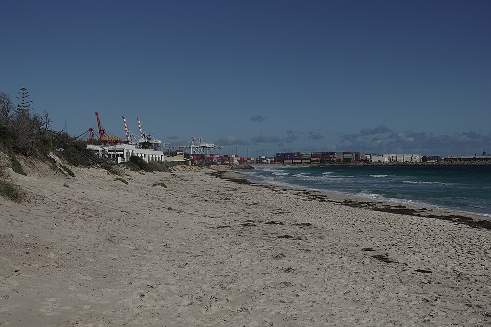 Sand and sea at Port Beach with port cranes and container stacks in the distance.