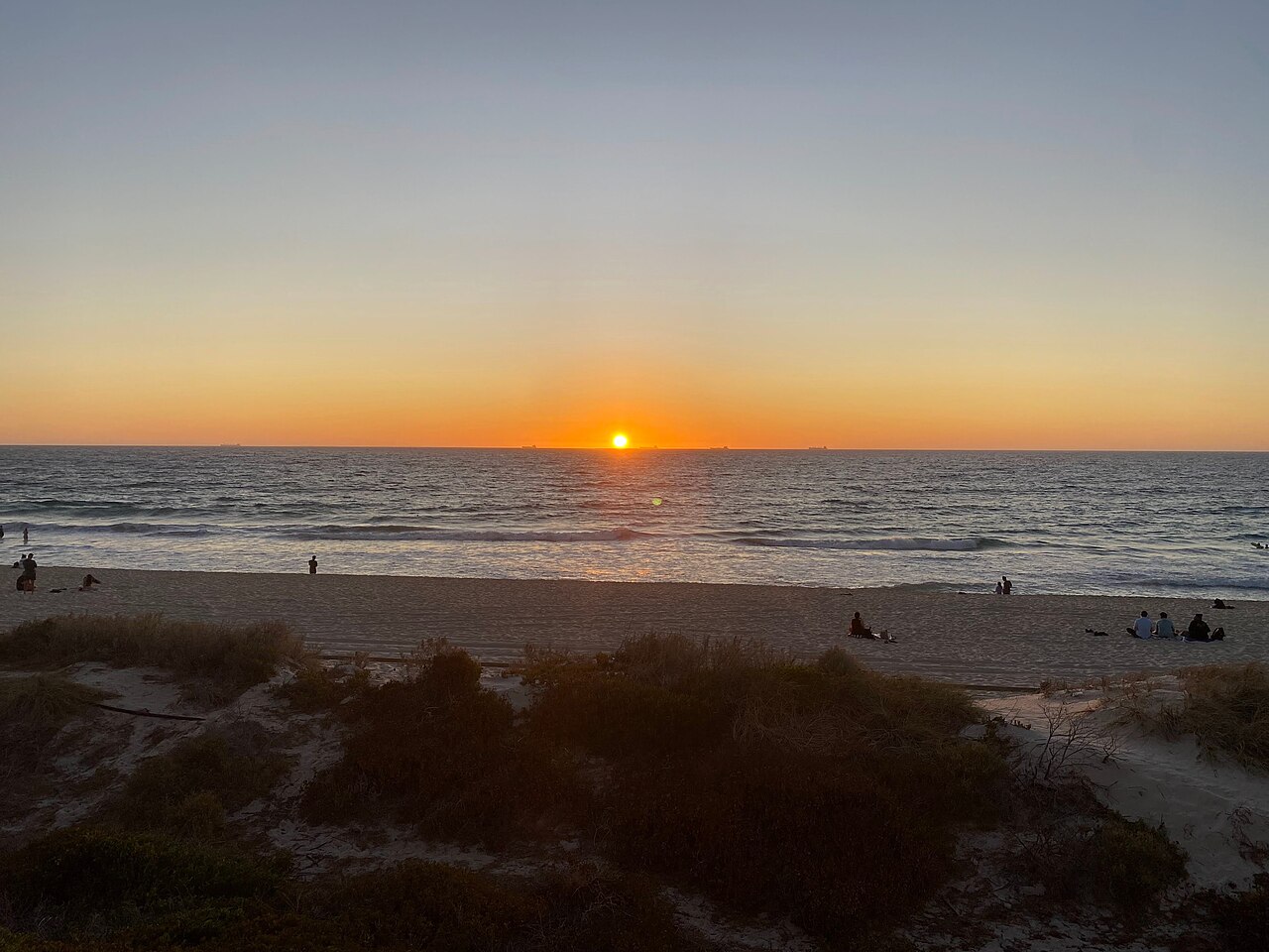 Sun setting over the Indian Ocean at Scarborough Beach, with sand and low coastal vegetation in the foreground.